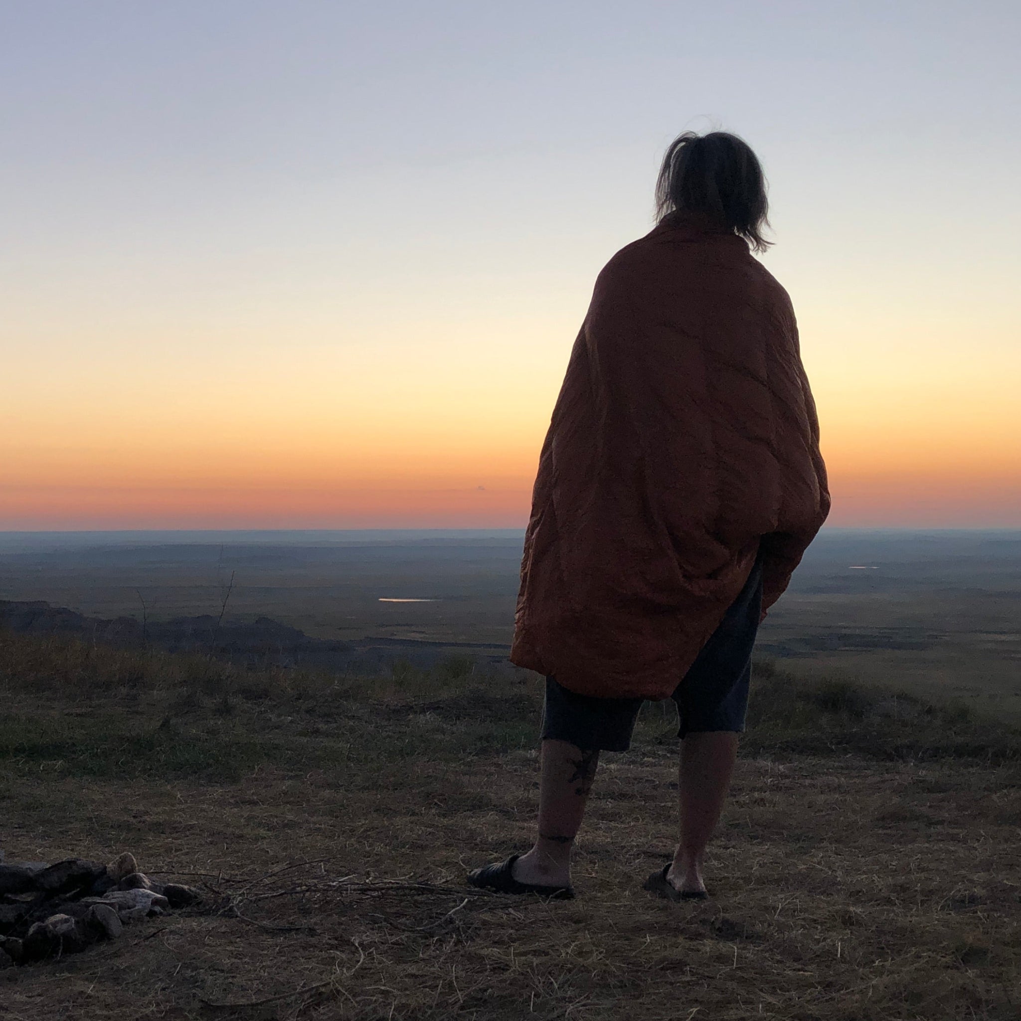 Camper wrapped in a Jacks R Better Shenandoah down camping quilt watching sunrise from a hilltop campsite